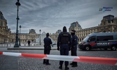 French police officers stand behind red and white caution tape in front of the Louvre Museum's pyramid and historical buildings in Paris, indicating a crime scene or security incident.