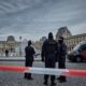 French police officers stand behind red and white caution tape in front of the Louvre Museum's pyramid and historical buildings in Paris, indicating a crime scene or security incident.
