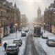 Pedestrians in winter coats walk down a frosty UK city street, with a light dusting of snow or sleet visible on rooftops and in a nearby park, illustrating the weekend cold snap.