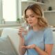 Young woman sitting on a sofa taking antidepressant medication with a glass of water, illustrating the importance of understanding physical health effects.